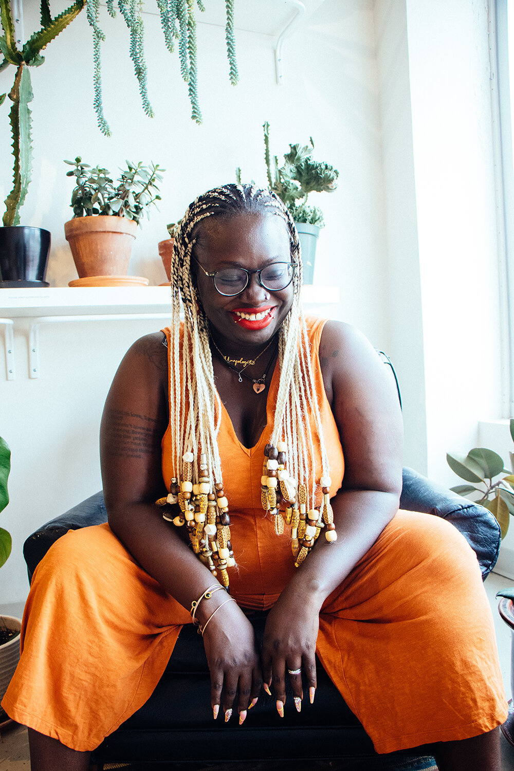 Brittany Rogers is sitting in a white room with potted plants behind her. She laughs towards the right corner while she rests her arms on her spread out legs.