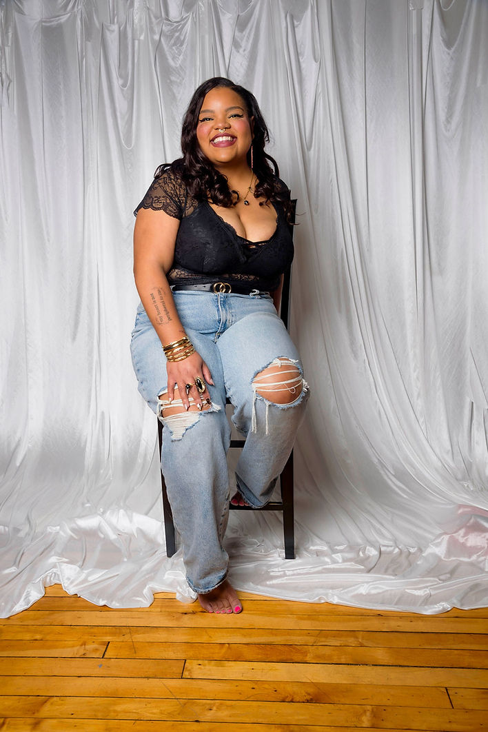 M. Mick Powell is sitting on a chair as they smile towards the camera. Their background is a white fabric hanging up top with the wooden floor showing past their feet.