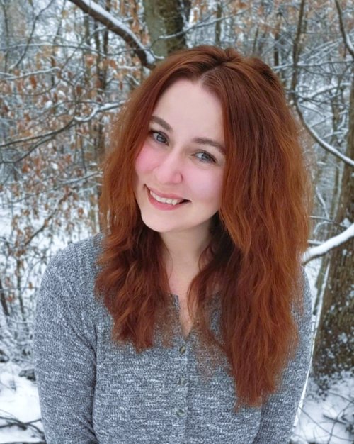 Raye Hendrix is smiling towards the camera with a bit of a head tilt. The background is snow outside with trees covered in little leaves covering the area.