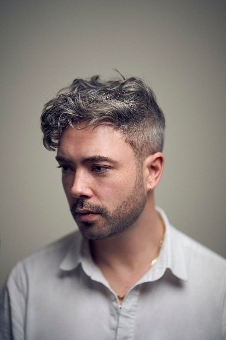Seán Hewitt is staring off into the distance of the left corner with a serious face. His gray background is faded along with his body from the neck down.