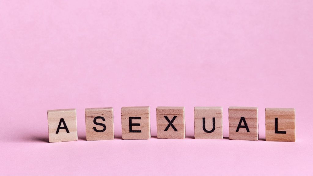 Scrabble tiles arranged upright to spell the word "asexual" on a light pink background.