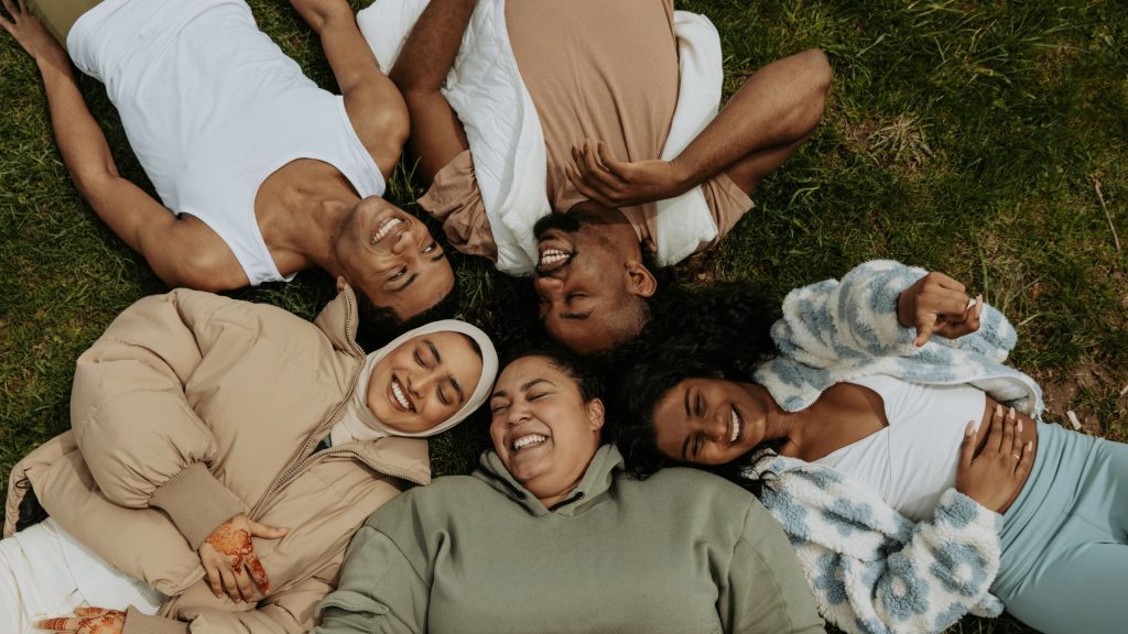 Five friends, all Black or Brown and dressed in neutral tones, lie smiling in the grass in a cluster with their heads facing inward.