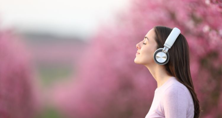 A woman with her eyes closed listening to headphones with a pink background.