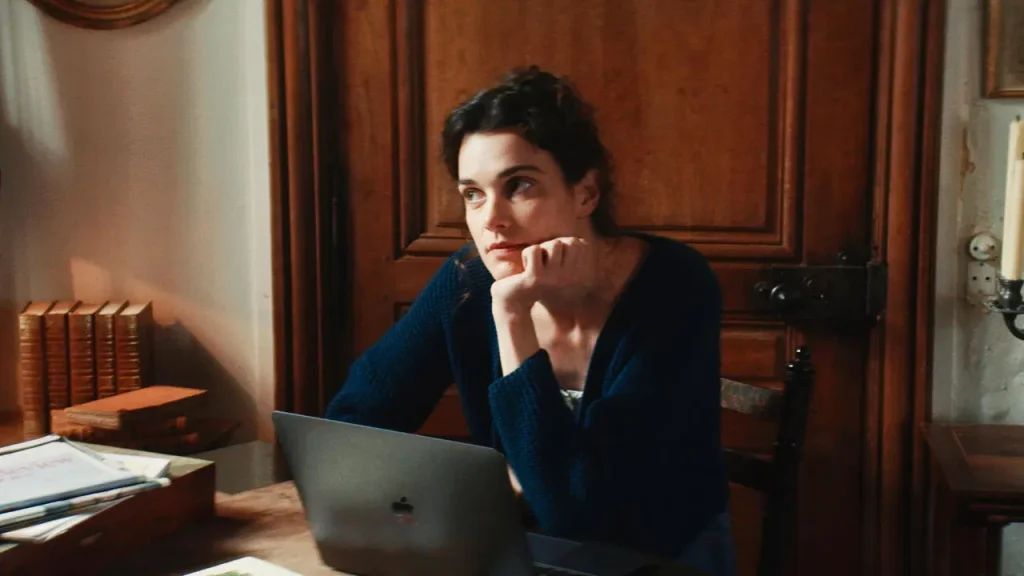 A still from Jane Austen Wrecked My Life. A woman in a blue sweater sits in front of a laptop with her chin on her hand. Beside her are antique leather bound books and writing notes. Behind her is a brown wooden door.