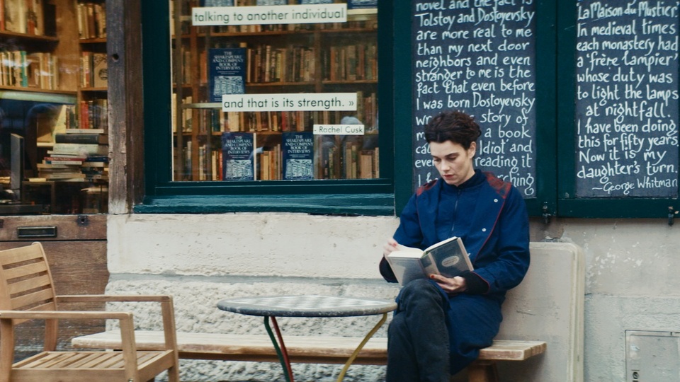A still from Jane Austen Wrecked My Life. A woman wearing a blue coat and jeans sits on a bench in front of a bookstore. The shop window behind her is covered in book quotes.