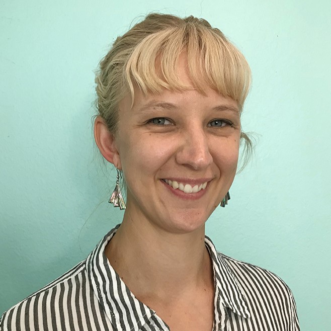 Lindsey Yankey, a smiling light-skinned woman with straight blonde hair tied back and bangs, as well as blue eyes. She wears a black-and-white vertically striped button down shirt and dangling silver earrings and stands in front of a light blue backdrop.
