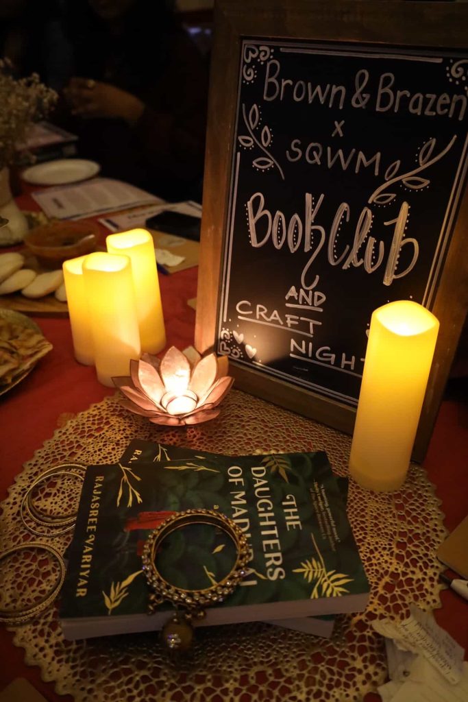A sign stating "Brown & Brazen x SQWM Book Club and Craft Night" surrounded by candles and a lotus-shaped diya. In front, are two stacked copies of The Daughters of Madurai by Rajasree Variyar with a gold bangle resting on top.