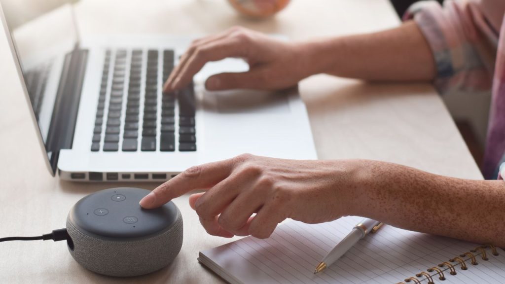 A person uses a laptop on a desk, touching a smart speaker button. A notebook with a pen lies nearby, conveying a tech-focused and productive atmosphere.