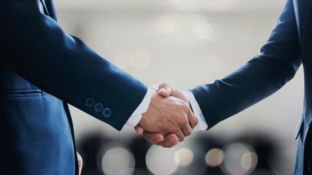 Close-up of two men in dark suits shaking hands, symbolizing agreement or partnership. The blurred background adds a professional tone.