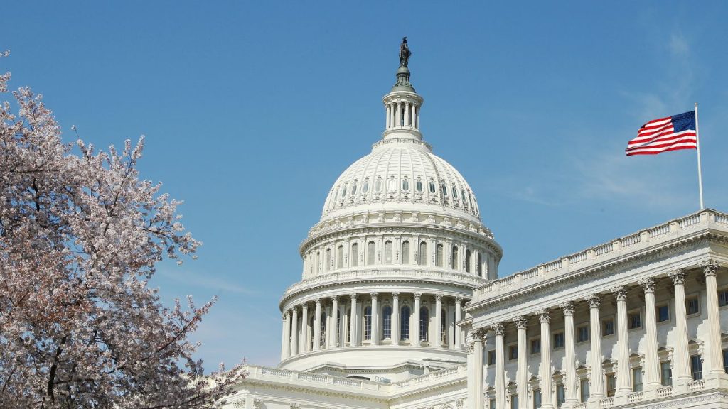 U.S. Capitol dome with a waving American flag on the right and blooming cherry blossoms on the left, set against a clear blue sky.