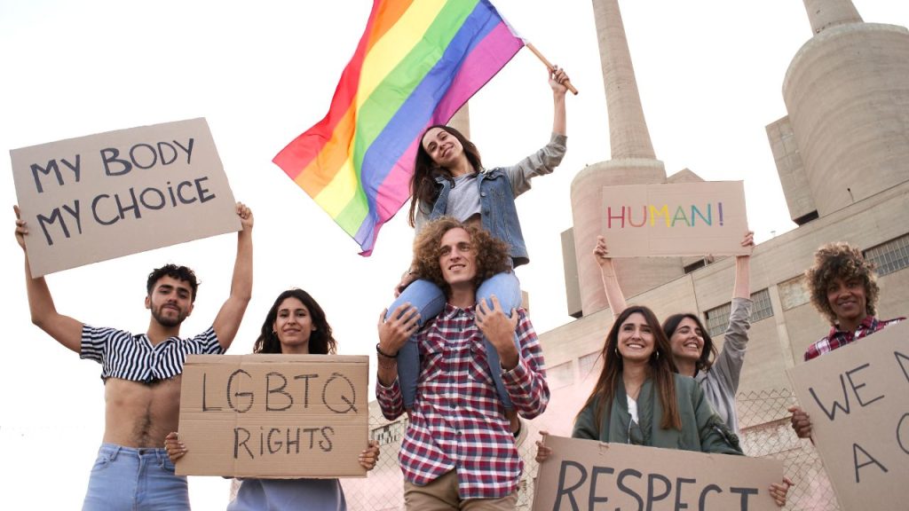 A diverse group of people hold signs for LGBTQ rights and equality. One person waves a rainbow flag. The mood is empowering and hopeful.