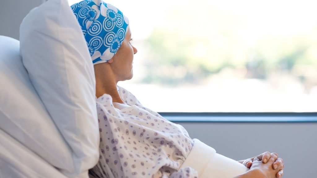 A person wearing a blue and white head covering and a white hospital gown lies in a hospital bed and looks out the window.