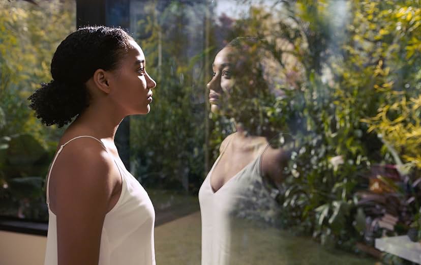 A still from Everything, Everything. A young Black girl wears a white tank top and stares out a large window into lush greenery. We can see her reflection in the glass.