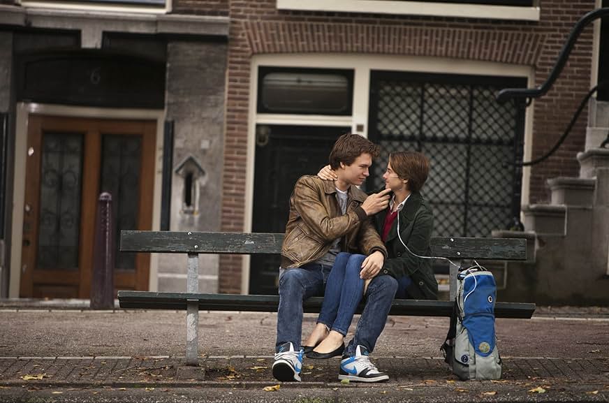 A still from The Fault in Our Stars. Two teens (a boy and a girl) sit on a park bench in front of a brick building. The boy wears a brown jacket and touches the girl's face. The girl wears a black jacket and red shirt and has an oxygen tube that goes to an oxygen tank tucked into a blue carrying case.