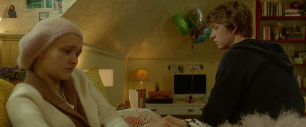 A still from Me and Earl and the Dying Girl. In a bedroom with white walls, a teen boy and girl sit on a bed, not looking at each other. The girl has a bald head and wears a pink beanie. She looks sad. Behind them are balloons and get well soon cards.