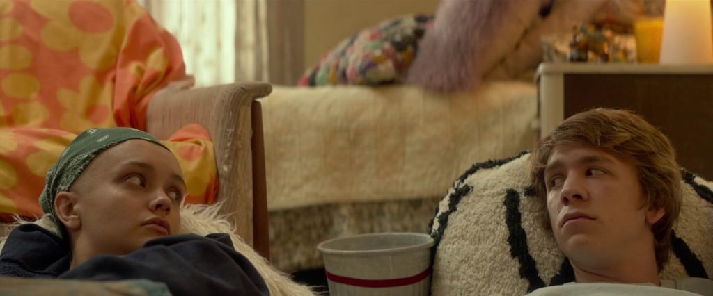 A still from Me and Earl and the Dying Girl. Two teenagers lean back on white cushions in a sunlit bedroom. There is an empty popcorn bucket between them. The girl on the left has a bald head and wears a green headscarf.