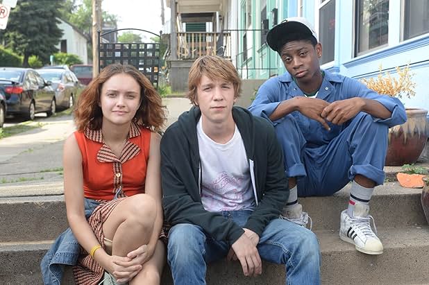 A still from Me and Earl and the Dying Girl. Three teenagers sit on sidewalk steps beside a blue house. On the left is a young white girl wearing an orange top. In the middle is a young white boy wearing a white t-shirt and black hoodie. On the right is a young Black boy wearing a blue jumpsuit and a white baseball cap.