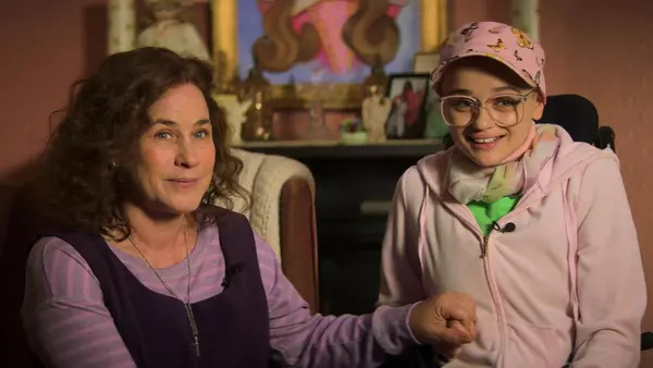 Scene from Hulu’s "The Act" featuring Patricia Arquette as Dee Dee Blanchard and Joey King as Gypsy Rose Blanchard. The two sit closely together, holding hands and smiling, talking to an interviewer. Dee Dee wears a purple vest over a striped shirt, while Gypsy, dressed in pink with a butterfly-printed cap and large glasses, appears frail and childlike.