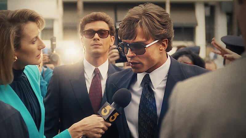 Nicholas Alexander Chavez as Lyle Menendez is interviewed by a reporter outside a courthouse, while Cooper Koch as Erik Menendez stands behind him in a suit and sunglasses. The scene is from "Monsters: The Lyle and Erik Menendez Story", a Netflix series created by Ryan Murphy.