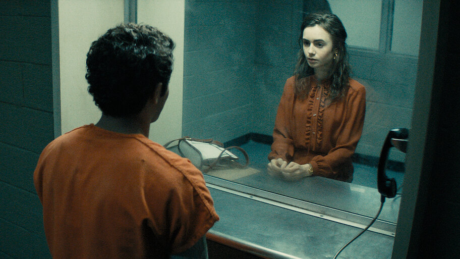 Scene from the film "Extremely Wicked, Shockingly Evil and Vile" showing Lily Collins as Liz Kendall visiting Zac Efron as Ted Bundy in prison. They are separated by a glass partition in a visitation room; Bundy wears an orange prison uniform while Liz sits seriously on the other side with a white purse next to her.