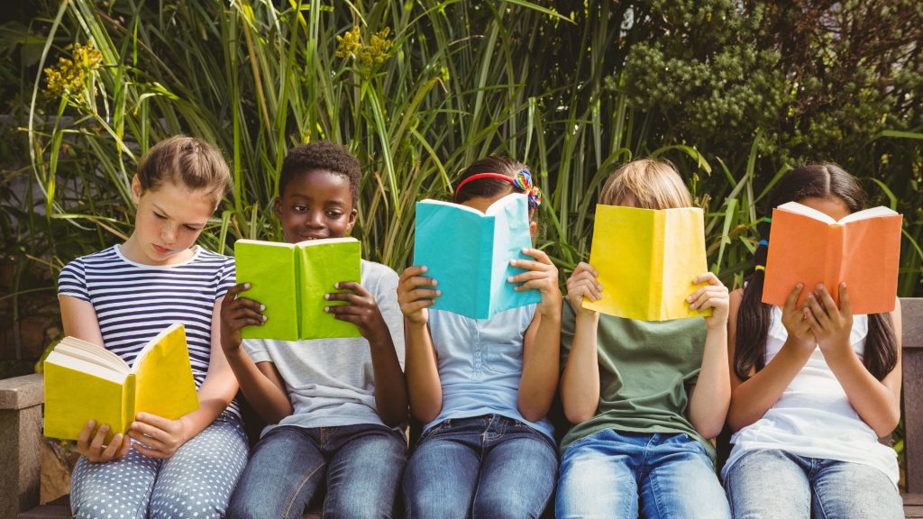 Five children sit side by side reading different-color books on a bench in front of some ferns.