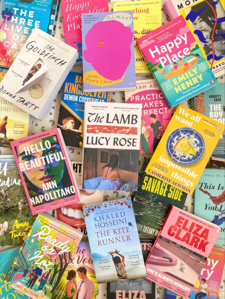 A colorful pile of fiction book covers. The top layer of book covers from top left to bottom right are The Goldfinch by Donna Tartt, Happy Place by Emily Henry, Hello Beautiful by Ann Napolitano, The Lamb by Lucy Rose, We All Want Impossible Things by Catherine Newman, and The Kite Runner by Khaled Hosseini.