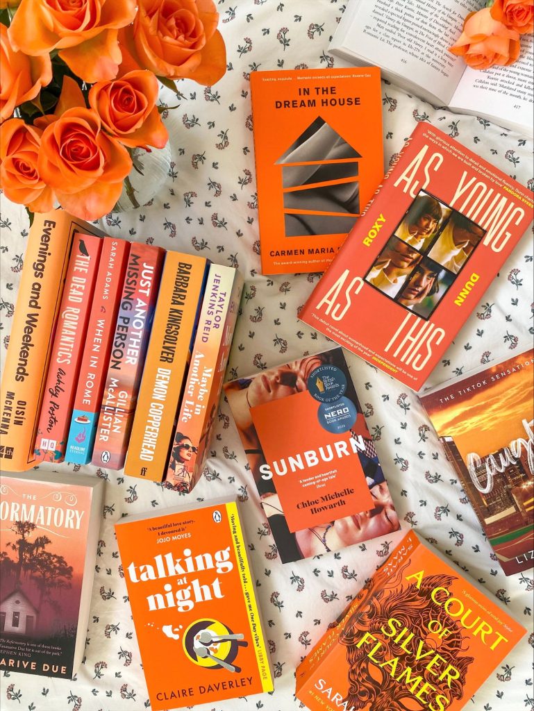 An array of orange fiction book covers on top of a white bedsheet speckled with little pink flowers.