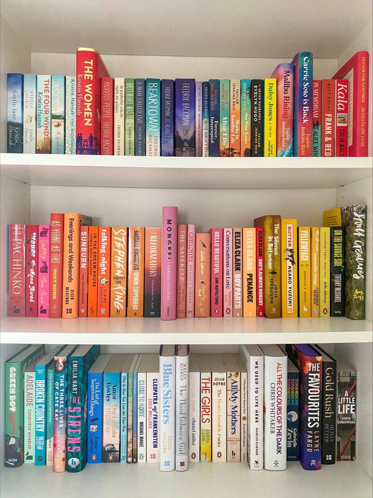 A white three-shelved bookcase full of colorful books. The top shelf has mainly blue and red shaded cook covers, the second shelft has arrays of pinks oranges, and yellows, and the bottom has light greens and blues into white.
