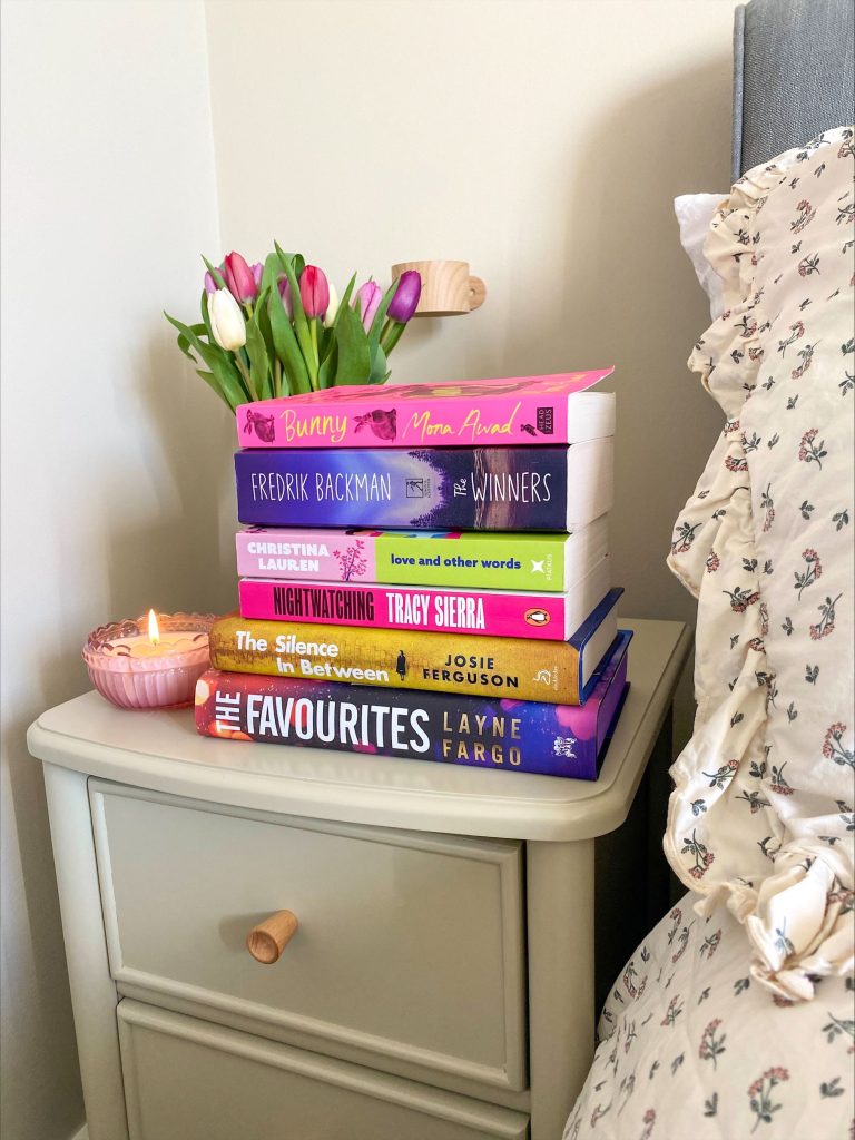 Bookstagrammer Lorna's night stand with a lit candle, pink and white tulips, and a stack of novels. From top to bottom the titles are Bunny by Mona Awad, The Winners by Fredrik Backman, Love and Other Words by Christina Lauren, Nightwatching by Tracy Sierra, The Silence In Between by Josie Ferguson, and The Favourites by Layne Fargo.