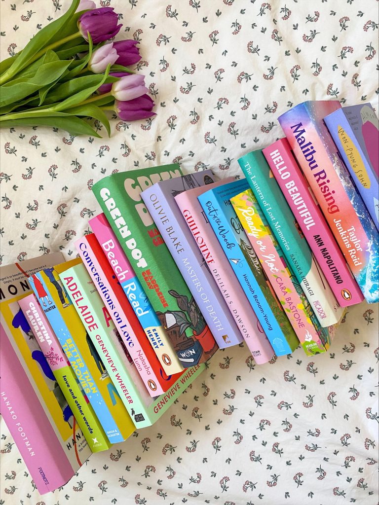 A pile of colorful books spilling over onto a white bedsheet with little pink flowers. In the corner is a loose bouquet of purple tulips.