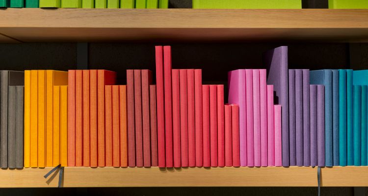 A brown bookshelf with rainbow coloured books on it (ranging from brown, yellow, orange, red, pink, purple, blue).