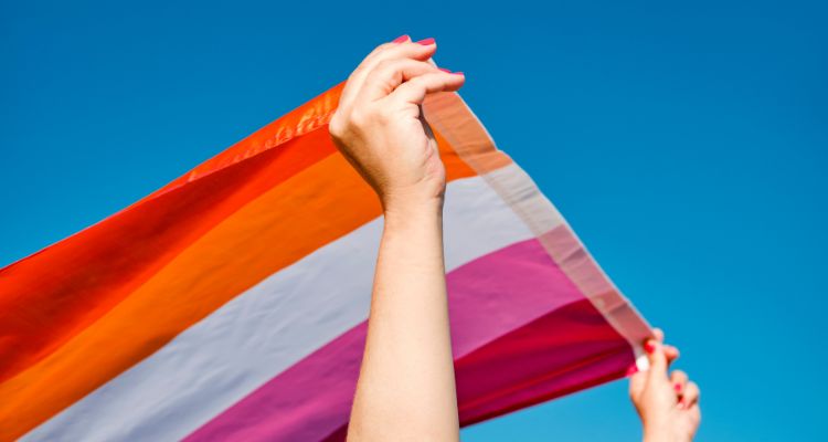 Person with pink painted fingernails holding a Lesbian flag up to a blue sky.