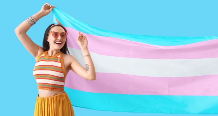 Person in orange crop top, skirt and love heart sunglasses holding the trans flag up in front of a light blue background.