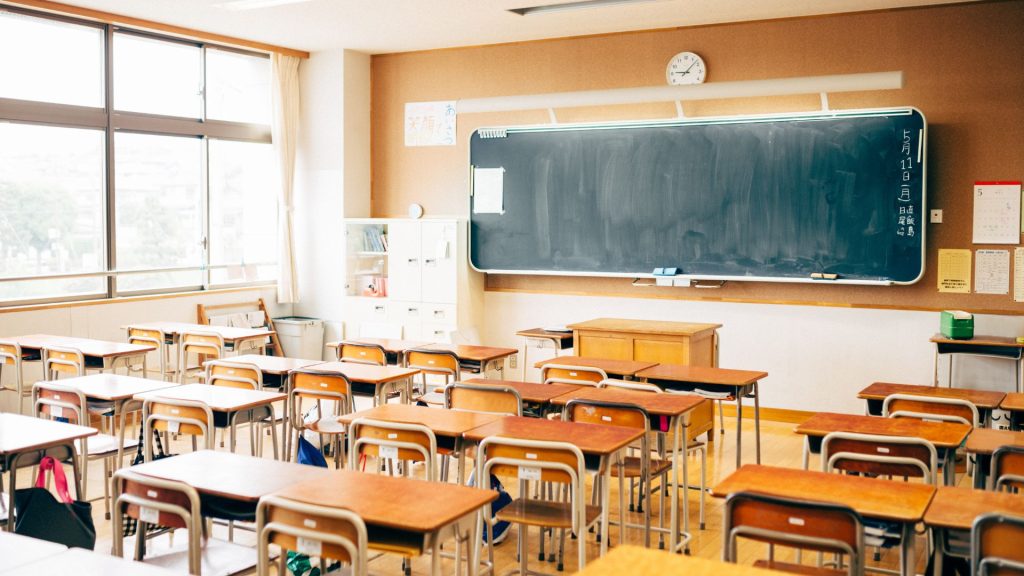 An empty classroom filled with wooden desks in sets of two, a blackboard with the date written on the side in Japanese, and a teacher's desk at the front.