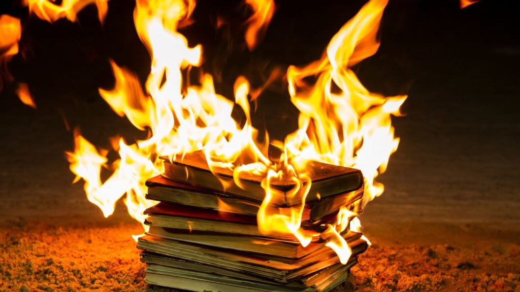 Stack of books engulfed in bright orange flames, burning on the ground in a dark setting, symbolizing destruction or censorship.