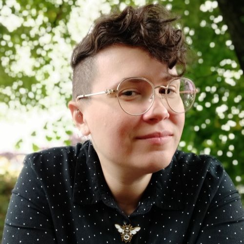 Niki Smith, a light-skinned woman with a curly brown undercut and light glasses, looks at the viewer with a tree in the background.