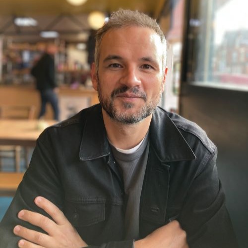 Anjan Sarkar looks at the viewer with his arms crossed in a cafe. He is a light-skinned man with gray hair and facial hair wearing a dark jacket and gray shirt, and a table, chairs, and counter with a patron approaching can be seen out of focus behind him.