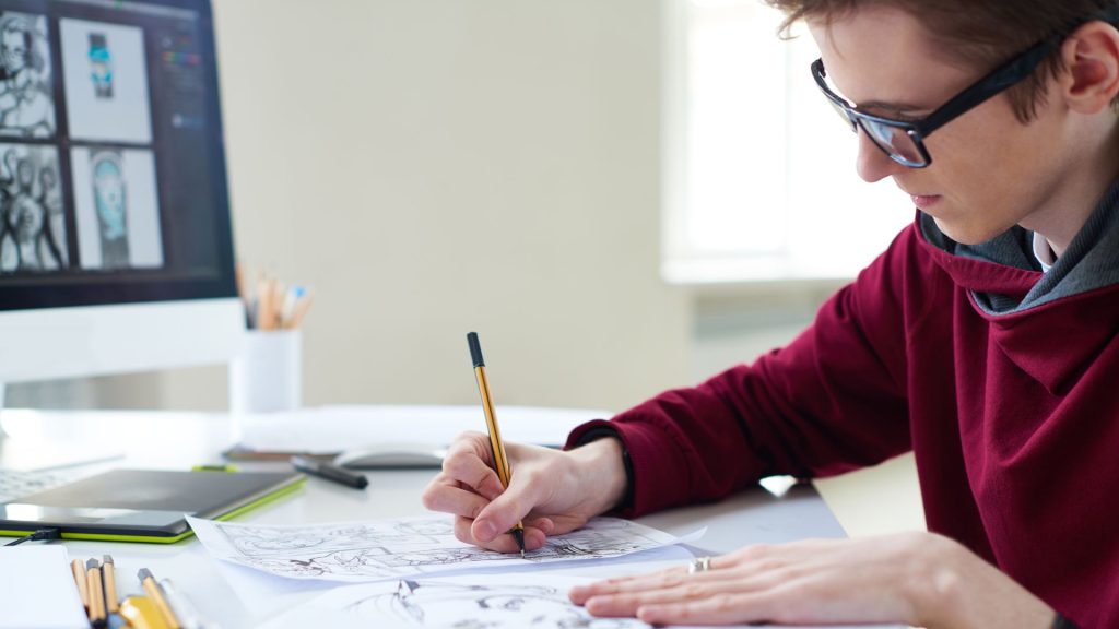 A light-skinned artist with short hair and glasses sits at a desk drawing a page for a graphic novel. On the desk are a mouse, tablet, computer monitor, and various pencils.