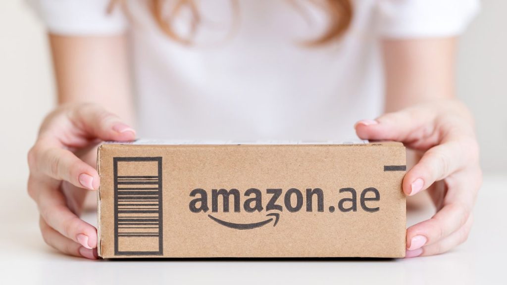 Hands holding a small cardboard box labeled "amazon.ae" with a barcode. The setting is minimalistic.