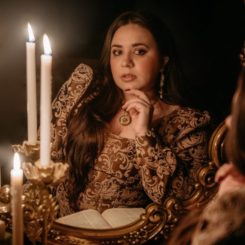 Kaylie Smith wears a brown brocade dress, a golden necklace, and golden earrings. She sits in front of a mirror in a swirling brown frame with an open book and candlesticks. She rests her chin on her hand and has a black background behind her.