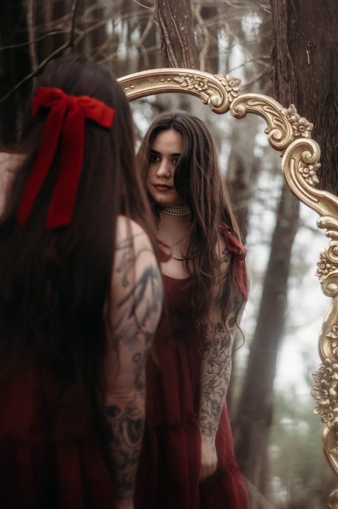 Kaylie Smith wears a red velvet dress, red hair bow, and pearl necklace. She stands in the woods in front of a mirror in a vintage gold frame. She has a full sleeve of tattoos.