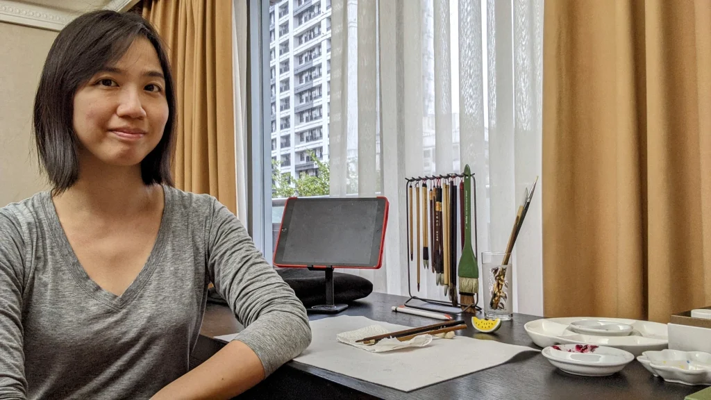 Regina Linke wears a gray shirt and sits at her art desk. There are painting supplies and a mounted tablet. In front of the desk is a window with white and golden brown curtains.