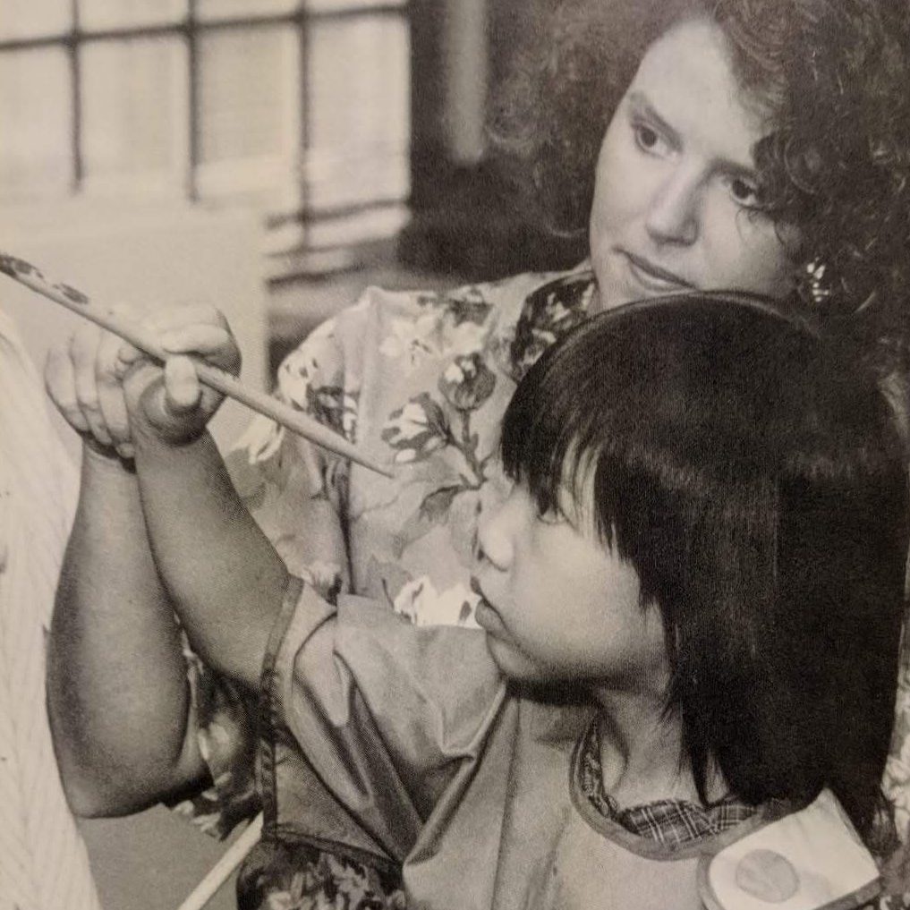 A black and white photo of a young Regina Linke being taught to paint by an older woman. They both wear art smocks. Regina holds a paintbrush against a canvas, and the woman guides her hand.