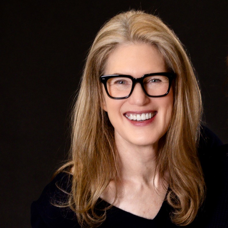 Sandra Nickel, a smiling light-skinned woman with long, straight blonde hair wearing black eyeglasses and a black blouse against a black background.