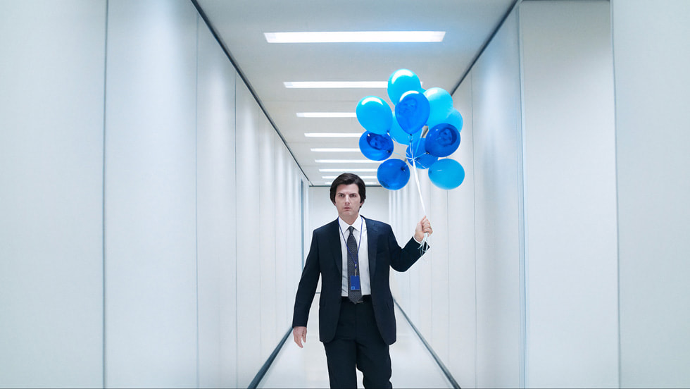 A still from the TV show Severance, featuring a light-skinned man with straight, short dark hair wearing a suit and holding a bunch of blue balloons, standing in a white hallway.
