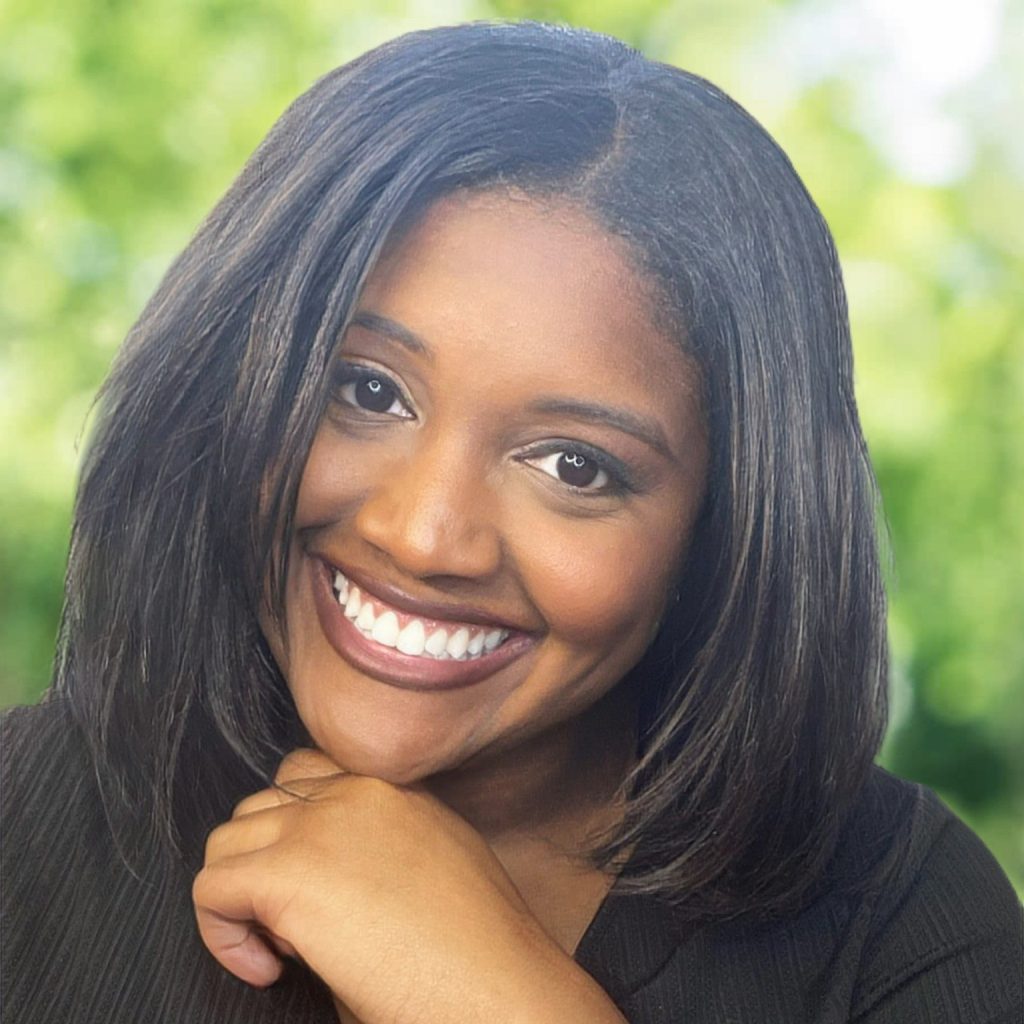 Gabi Burton, a Black woman with a straight bob hairstyle and black T-shirt, smiles at the viewer as she rests her chin on her hand. Out of focus in the background are some trees.