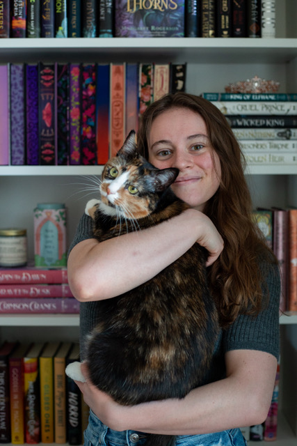 Author Isabella Stofka stands in front of bookshelves, holding her cat and smiling