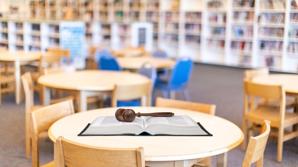 An open book with a gavel laying on top on a round table in a library with blurred bookshelves in the background.