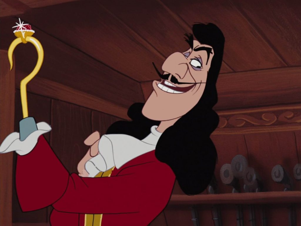 A still from Peter Pan (1953). Captain Hook wears a red coat and raises his gold hook hand with a smug expression on his face. Behind him is the wooden interior of a ship.