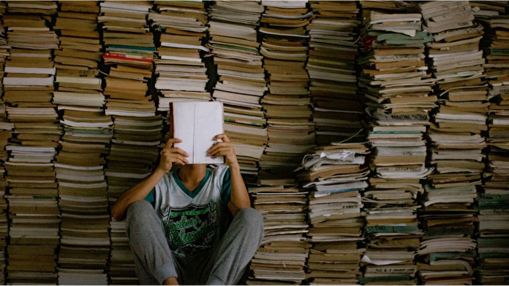 A person sits on the floor in front of a massive stack of books and loose papers, holding an open book in front of their face.